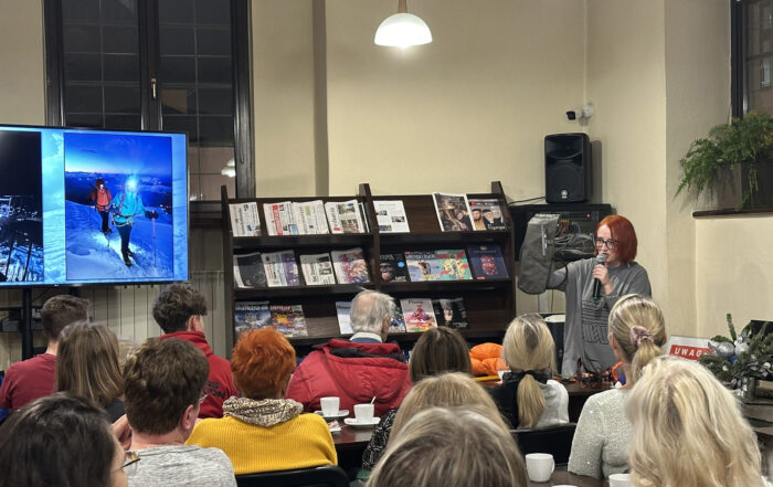 Kobieta z mikrofonem mówi do grupy ludzi. W tle widać duży ekran ze zdjęciami i rozłożone gazety na półkach.