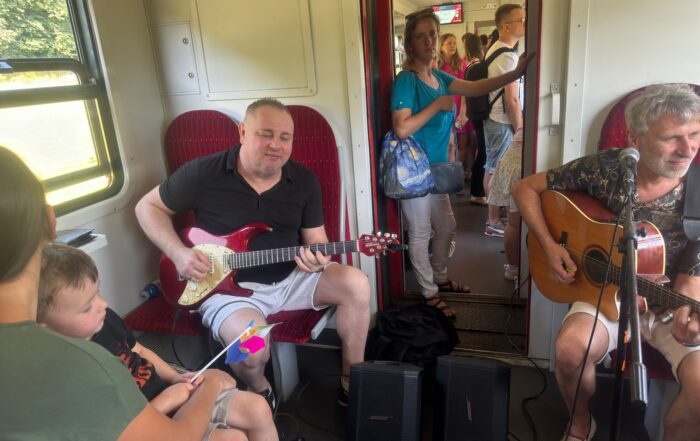 Dwóch mężczyzn siedzi na fotelach w pociągu. Grają na gitarach. Pomiędzy nimi w przejściu stoją dwa głośniki. Na przeciwko jednego z nich po lewej stronie siedzi kobieta z dzieckiem na kolanach. Chłopiec trzyma w rękach mały kolorowy wiatraczek. W tle widać przejście i kolejny przedział pełen ludzi. W przejściu stoi kobieta w średnim wieku w niebieskiej koszulce, białych spodniach, torebce i okularach we włosach.