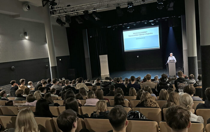 Aula w Akademii Nauk Stosowanych. W sali liczna grupa młodych ludzi, pokazana od tyłu, w rzędach siedzi na krzesłach. Przed nimi na scenie za mównicą stoi młody mężczyzna i mówi do mikrofonu. Za sceną w tle wisi wielki ekran. Na ekranie wyświetlone jest zdjęcie.