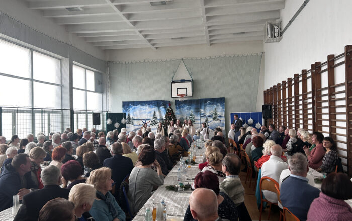 Tłum ludzi siedzi przy stołach i patrzy na śpiewające dzieci. Za dziećmi na ścianie wisi duży obraz z zimowym krajobrazem.