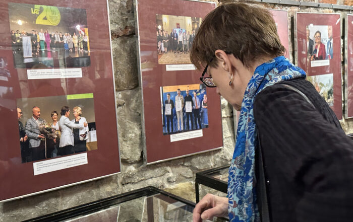 Kobieta w niebieskim szalu patrzy w dół. Przed nią wiszą kolorowe zdjęcia w ramach na bordowym tle. Na zdjęciach grupy ludzi.