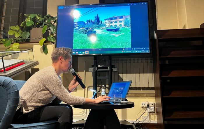 Kobieta w szarym swetrze siedzi na fotelu i pochyla się nad niskim okrągłym stolikiem. Trzyma przy twarzy mikrofon . Na stoliku szklanka, butelka wody i otwarty laptop. Nad nią duży ekran ze zdjęciem.