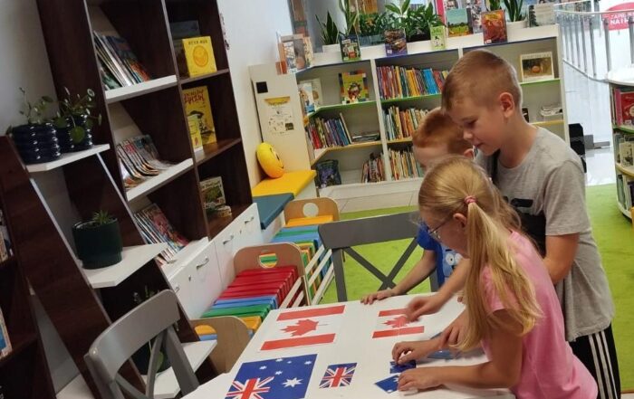 Troje dzieci układa flagi państw w sali na małym białym stoliku. Na stole flaga kanady i Nowej Zelandii. W tle regały z kolorowymi książeczkami i półki z doniczkami z kwiatami.