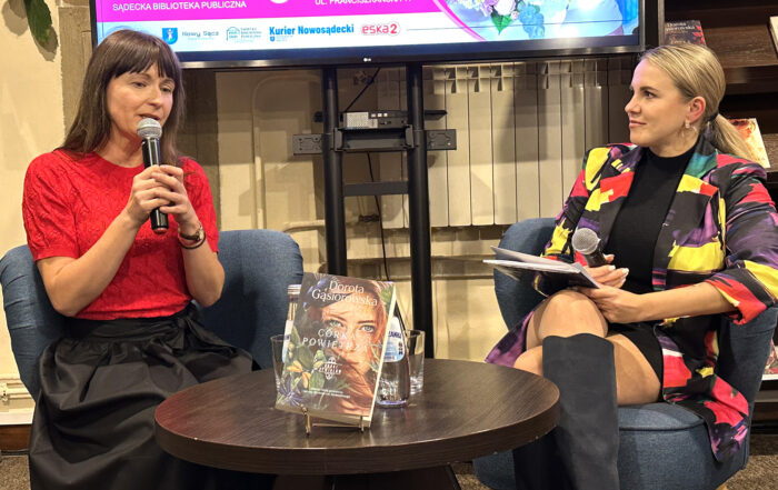 Dwie kobiety w sali. Siedzą na fotelach przy małym okrągłym stoliku. Na stoliku książka, szklanki i dwie butelki wody. Jedna z nich ma dłuższe brązowe włosy, czerwoną bluzkę i czarną długą spódnicę. Trzyma przy twarzy mikrofon. Druga z kobiet ma wielokolorową marynarkę i długie ciemne kozaki. Patrzy z zaciekawieniem na kobietę w czerwonej bluzce. Za nimi fragment ekranu.