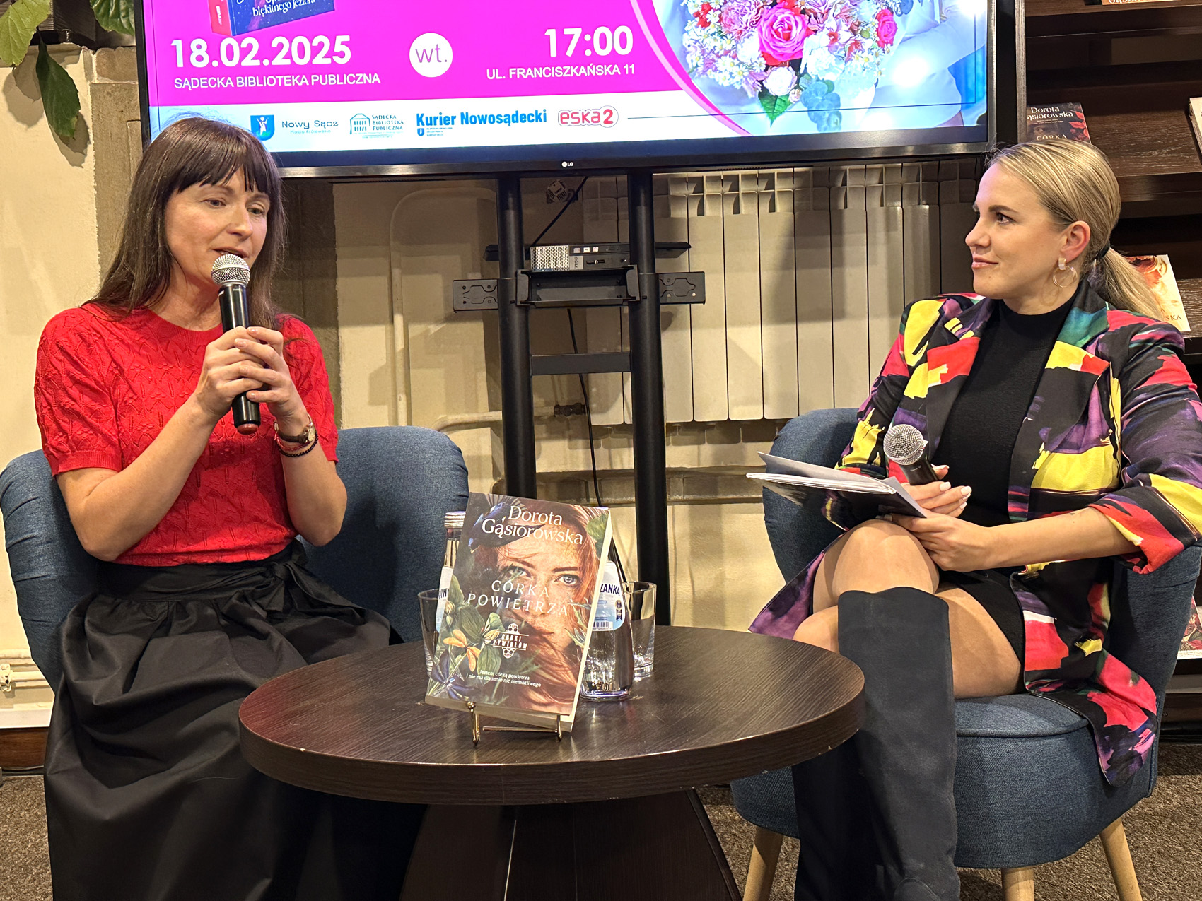 Dwie kobiety w sali. Siedzą na fotelach przy małym okrągłym stoliku. Na stoliku książka, szklanki i dwie butelki wody. Jedna z nich ma dłuższe brązowe włosy, czerwoną bluzkę i czarną długą spódnicę. Trzyma przy twarzy mikrofon. Druga z kobiet ma wielokolorową marynarkę i długie ciemne kozaki. Patrzy z zaciekawieniem na kobietę w czerwonej bluzce. Za nimi fragment ekranu.