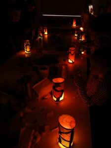 Ludzie w pomieszczeniu siedzą przy stole przy zapalonych lampionach. Bardzo ciemne wnętrze.