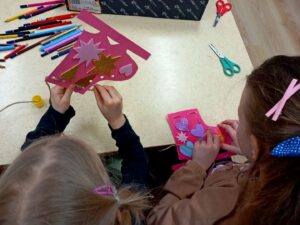 Dzieci robiące kolorowe ozdoby. Przed nimi stół. Na stole dużo kredek, klej i nożyczki.