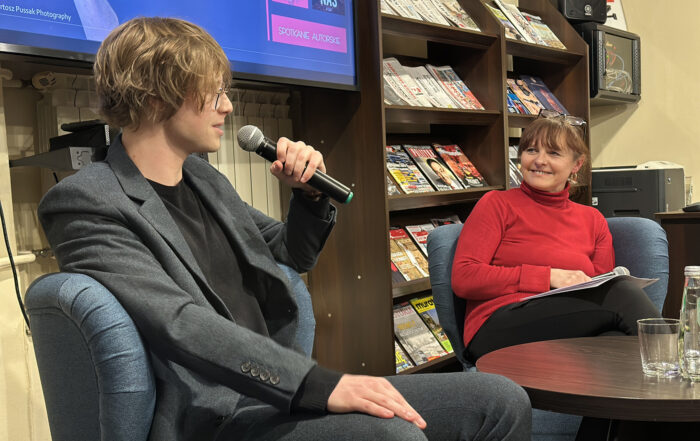 Młody mężczyzna w garniturze trzyma mikrofon w ręce. Kobieta w czerwonej bluzce patrzy w jego kierunku. Ma okulary założone we włosach. Uśmiecha się. Za nią regal z gazetami.