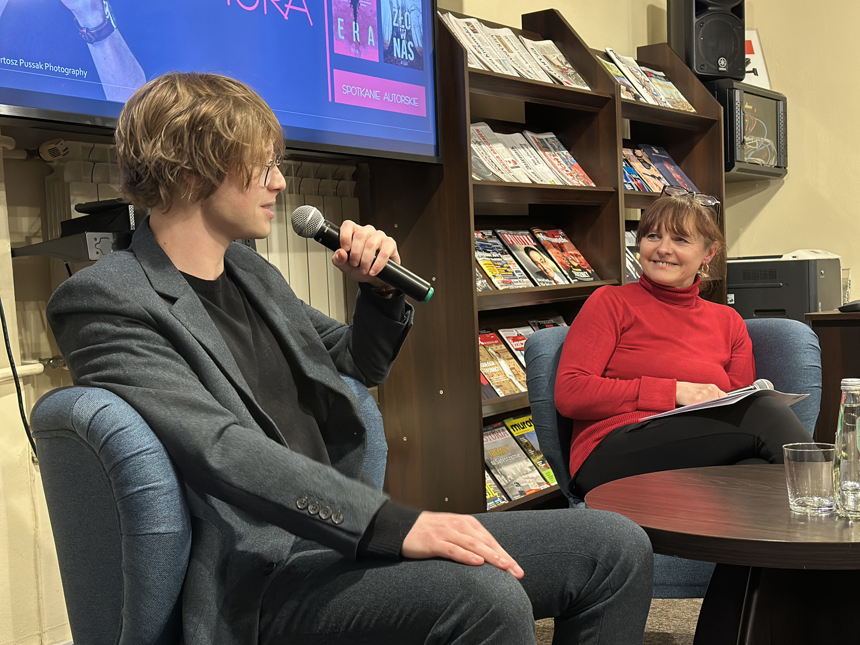 Młody mężczyzna w garniturze trzyma mikrofon w ręce. Kobieta w czerwonej bluzce patrzy w jego kierunku. Ma okulary założone we włosach. Uśmiecha się. Za nią regal z gazetami.