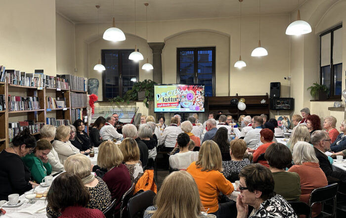 Kilkadziesiąt osób siedzi przy stołach w sali. Na stołach są białe obrusy, szklanki i pączki na talerzach. Na froncie ekran z kolorową grafiką. Na grafice postać w masce karnawałowej. Po lewej stronie regały z książkami. Z sufitu zwisają zapalone lampy w kloszach.