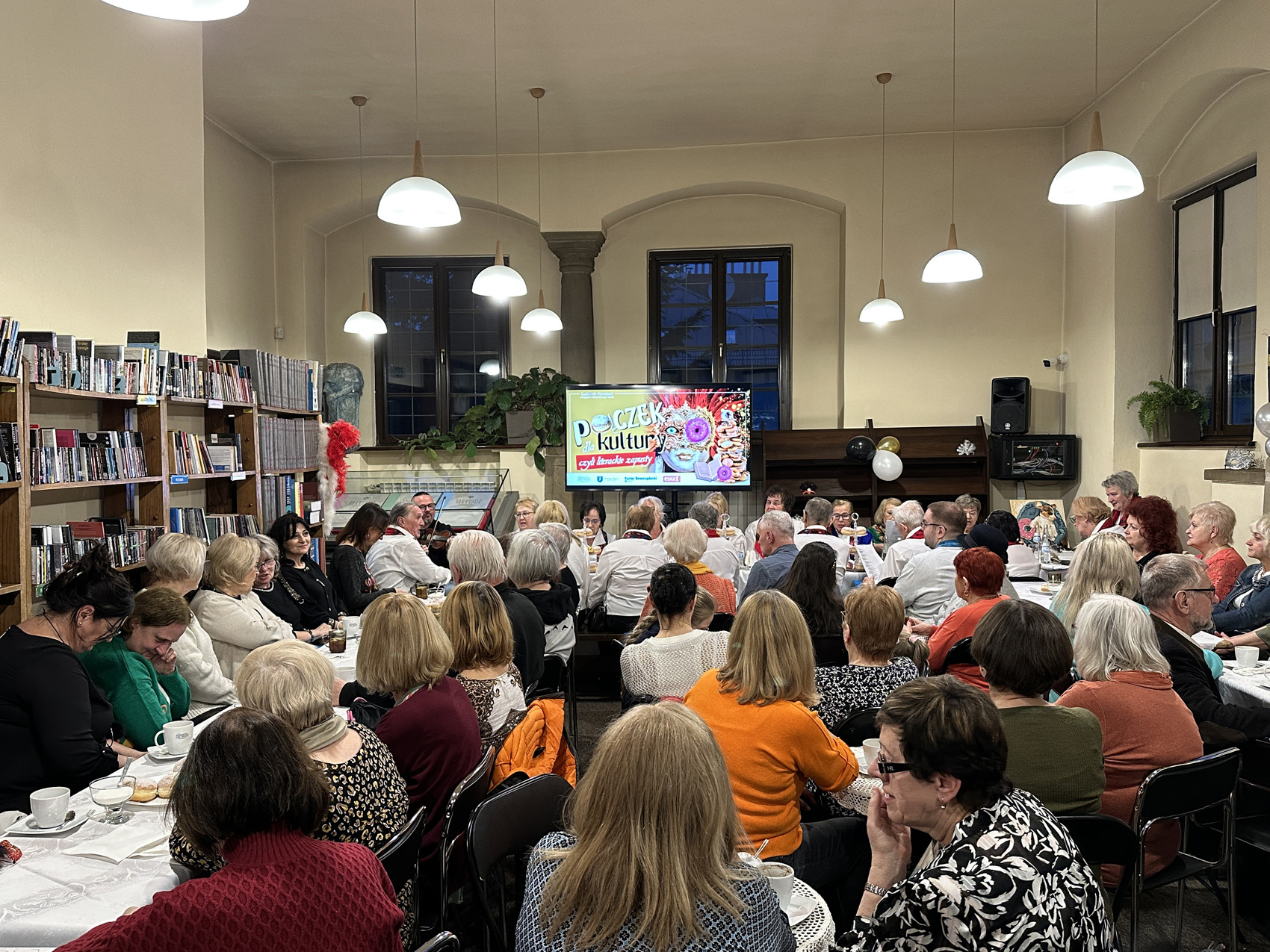 Kilkadziesiąt osób siedzi przy stołach w sali. Na stołach są białe obrusy, szklanki i pączki na talerzach. Na froncie ekran z kolorową grafiką. Na grafice postać w masce karnawałowej. Po lewej stronie regały z książkami. Z sufitu zwisają zapalone lampy w kloszach.