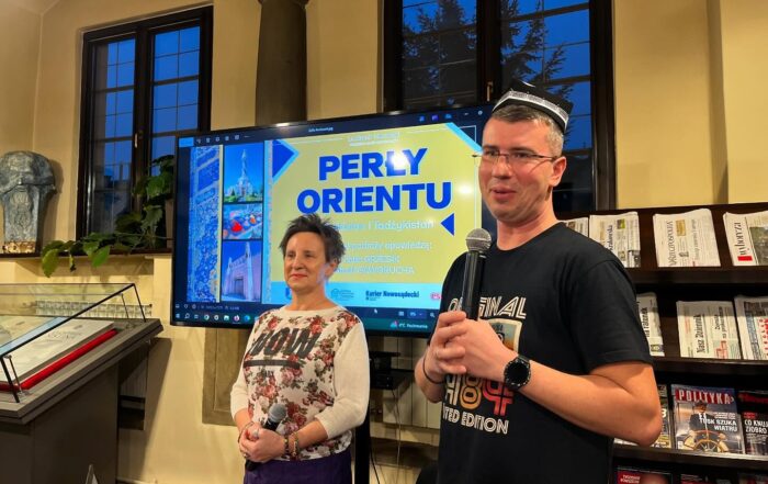 Mężczyzna w czapce i koszulce trzyma mikrofon. Za nim znajduje się duży ekran z kolorową grafiką: „PERŁY ORIENTU”. Uśmiecha się. Obok niego stoi kobieta w średnim wieku uśmiecha się. Za nimi duże okna.