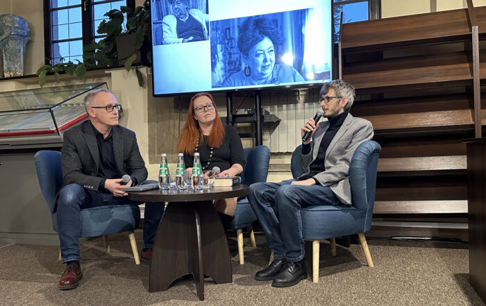 Dwaj mężczyźni w okularach i garniturach i jedna kobieta w czarnej sukience siedzą przy małym okrągłym stoliku. Na nim szklane butelki z wodą i szklanki. Za nimi w tle duży ekran z czarno-białymi zdjęciami.