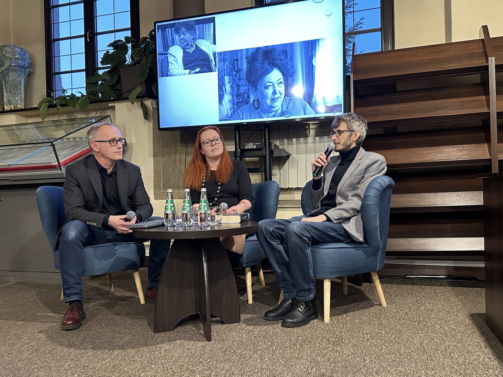 Dwaj mężczyźni w okularach i garniturach i jedna kobieta w czarnej sukience siedzą przy małym okrągłym stoliku. Na nim szklane butelki z wodą i szklanki. Za nimi w tle duży ekran z czarno-białymi zdjęciami.