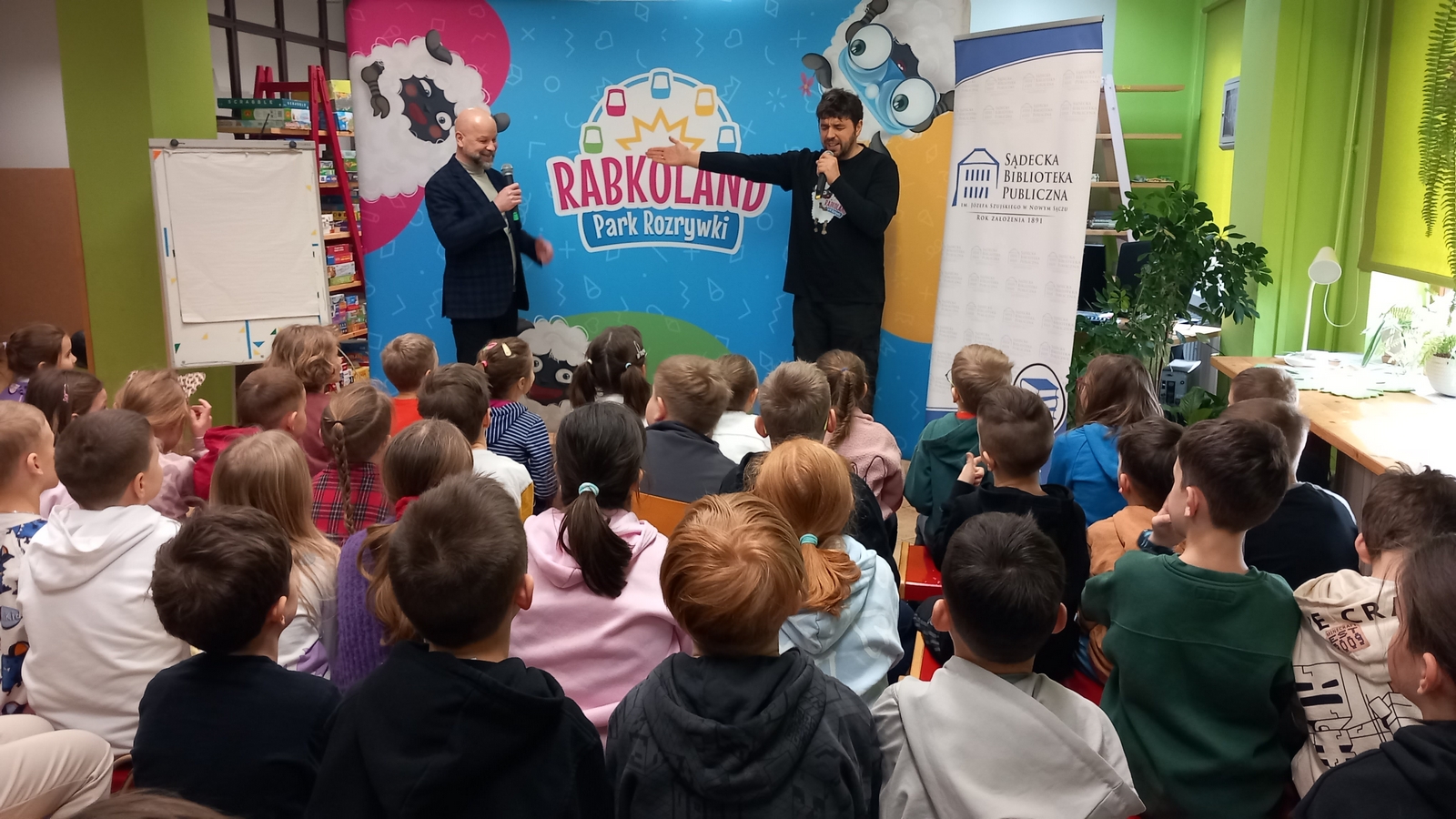 Młody mężczyzna w ciemnych kręconych włosach stoi na tle kolorowej ścinaki z owcami. Pokazuje ręką na mężczyznę obok. Drugi z mężczyzn jest łysy. uśmiecha się. Ubrany jest w ciemny garnitur i trzyma mikrofon w ręce. Obok stoi baner Sądeckiej Biblioteki Publicznej. Przed nim znajduje się duża grupa dzieci.