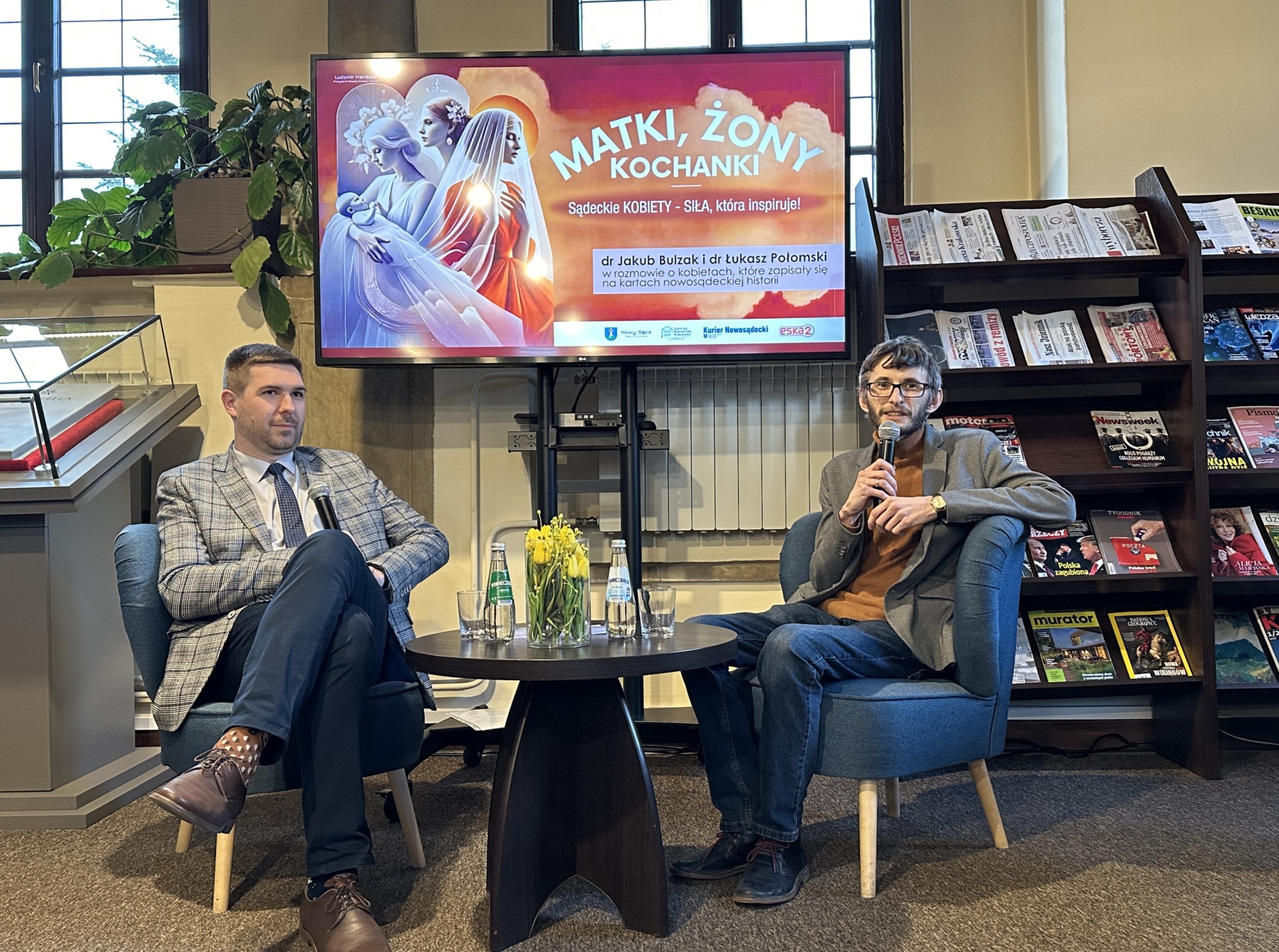 Dwóch mężczyzn siedzi przy okrągłym brązowym stoliku. Jeden z nich ma szarą marynarkę w kratę oraz granatowe spodnie. Drugi okulary, czarną krótka brodę i ma na sobie brązowy golf i szarą marynarkę. Na stole szklany flakon z żółtymi tulipanami, dwie szklanki i dwie szklane butelki z wodą mineralną. W tle duży ekran z kolorową grafiką oraz regał z gazetami i kolorowymi magazynami.