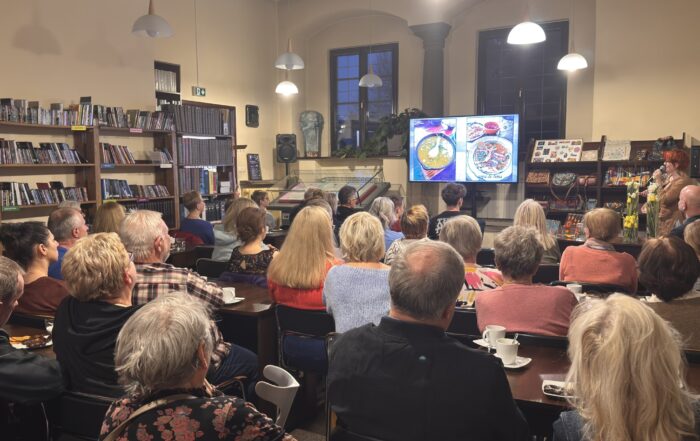 Grupa ludzi na sali siedzi na krzesłach i patrzy na front sali. Z przodu kobieta w krótkich rudych włosach mówi do mikrofonu. Obok niej jest regał z kolorowymi magnesami i torebkami. Na froncie duży telewizor. Na ekranie wyświetlają się zdjęcia. Po lewej stronie stoją regały z książkami.