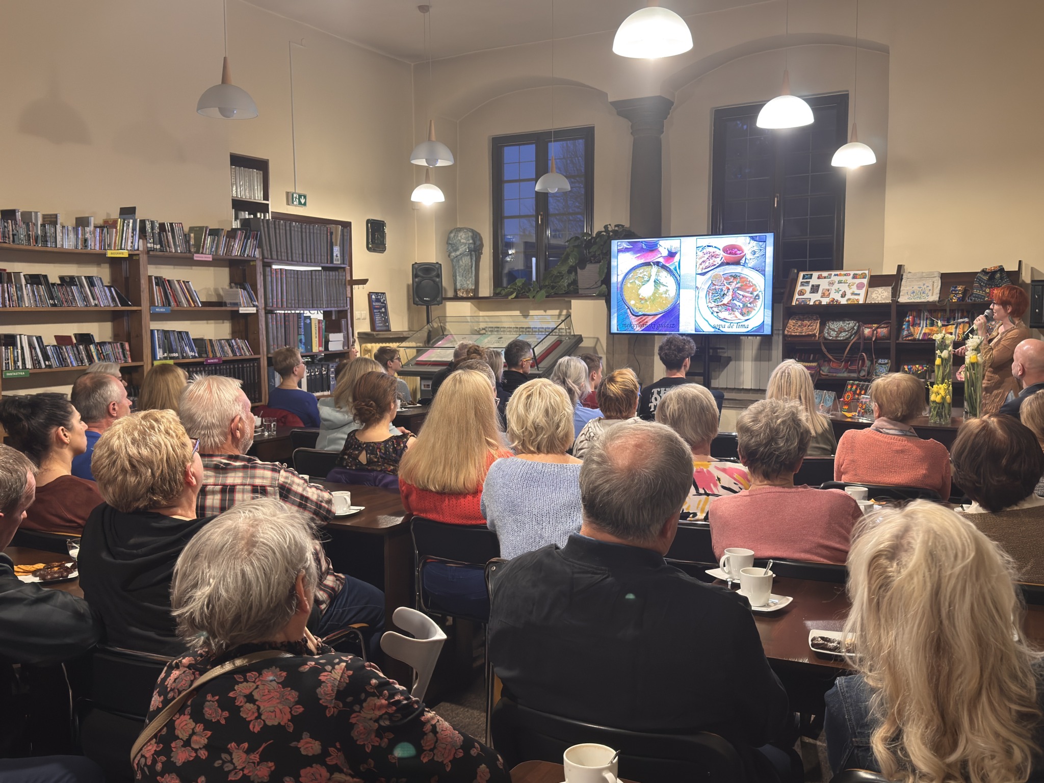 Grupa ludzi na sali siedzi na krzesłach i patrzy na front sali. Z przodu kobieta w krótkich rudych włosach mówi do mikrofonu. Obok niej jest regał z kolorowymi magnesami i torebkami. Na froncie duży telewizor. Na ekranie wyświetlają się zdjęcia. Po lewej stronie stoją regały z książkami.