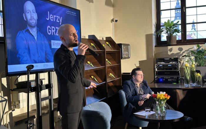 Mężczyzna w czarnym garniturze śpiewa z zamkniętymi oczami do mikrofonu. Za nim duży ekran z kolorowa grafiką. Obok przy brązowym okrągłym stoliku siedzi brunet w średnim wieku w czarnym garniturze i biało czarnej koszuli w geometryczne wzory. Na stole flakon z żółtymi tulipanami.