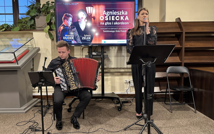 Dwoje ludzi w sali. Mężczyzna po lewej siedzi na krześle. Jest ubrany cały na czarno. Gra na czarno-złoto-czerwonym akordeonie. Patrzy przed siebie na tablet. Tablet stoi na czarnym stojaku. Obok niego stoi kobieta w czarnych spodniach i czarnej koronkowej bluzce. Ma długie jasnobrązowe włosy. Trzyma ręce na stojaku i mikrofonie. Jest bardzo skupiona. Przed nią stoi czarna tacka. Za nimi duży ekran z kolorową grafiką, duże okna i ciemno-brązowy regał.