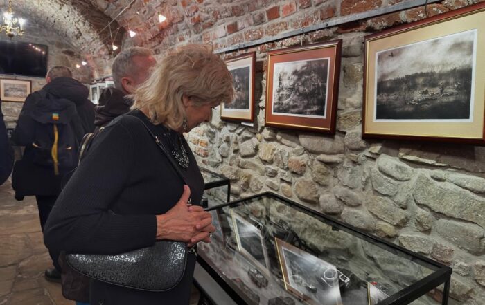 Ludzie w pomieszczeniu z ceglanymi ścianami. Na ścianach wiszą biało-czarne obrazy w brązowych ramkach. Pod nimi są szklane gabloty. W gablotach są aparaty fotograficzne. Na pierwszym planie jest kobieta w czarnej sukience, dużej błyszczącej biżuterii. Trzyma pod ramię czarną torebkę. Patrzy w dół na gabloty.