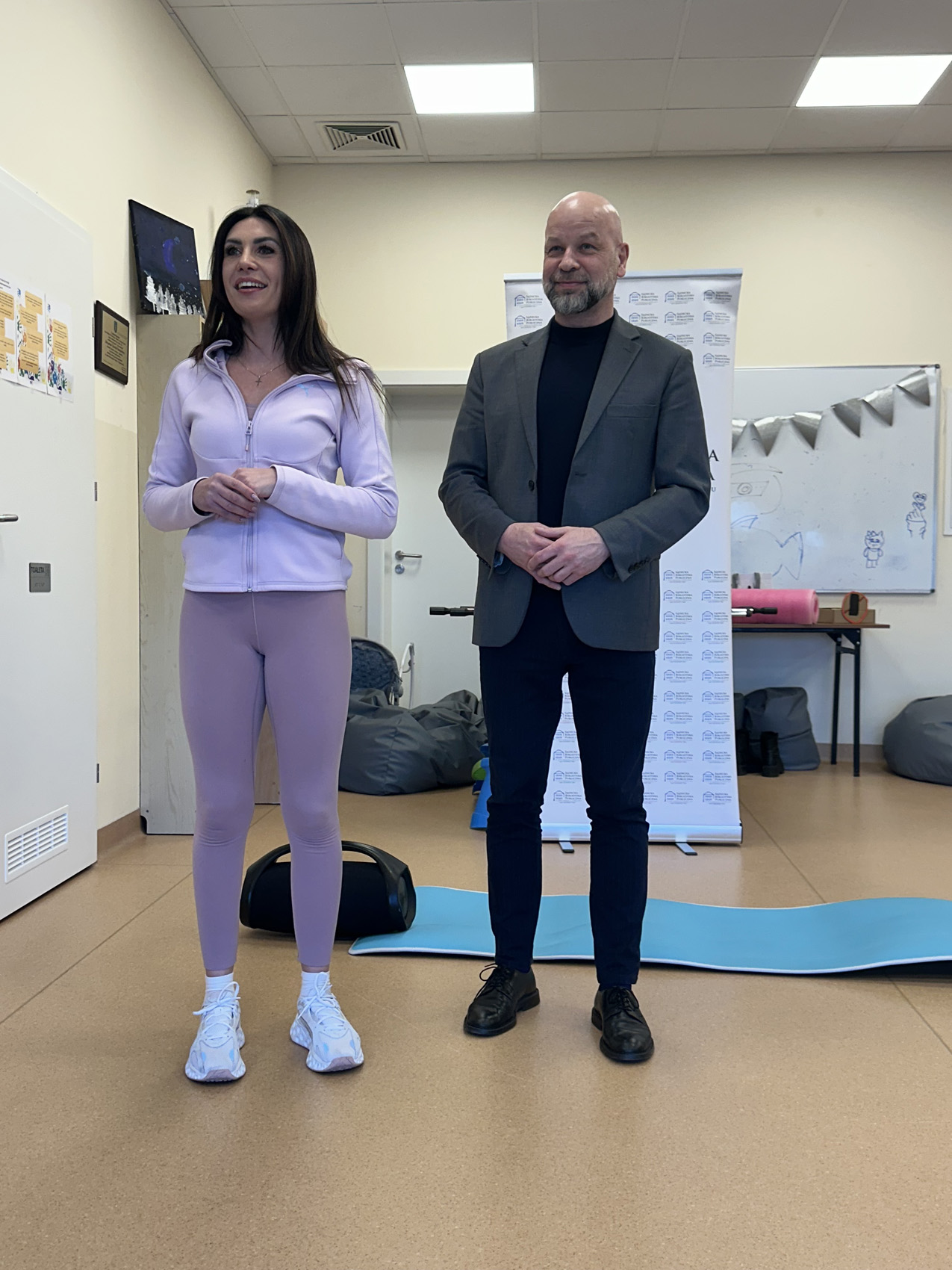Zajęcia ogólnorozwojowe 1a Kobieta i mężczyzna. Po lewej stronie stoi uśmiechnięta brunetka w jasnofioletowej bluzie sportowej i leginsach. Ma długie ciemne rozpuszczone włosy. Po prawej stoi uśmiechnięty łysy mężczyzna z krótką brodą w czarnych spodniach, czarnym półgolfie i ciemnoszarej marynarce. Za nimi baner i tablica. Na podłodze duży głośnik i niebieska mata do ćwiczeń.