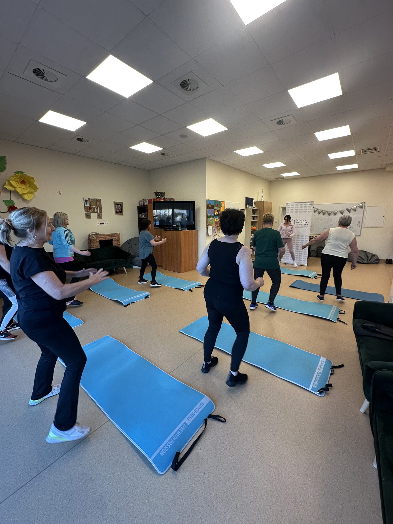 Zajęcia ogólnorozwojowe 2aa Osiem kobiet w sali. Przed nimi na podłodze niebieskie maty do ćwiczeń. Kobiety w strojach sportowych.