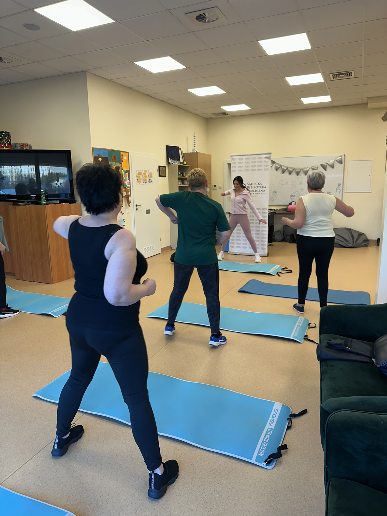 Zajęcia ogólnorozwojowe 4aa Cztery kobiety w sali. Przed nimi na podłodze niebieskie maty do ćwiczeń. Kobiety w strojach sportowych stoją w rozkroku. Mają podniesione ręce.