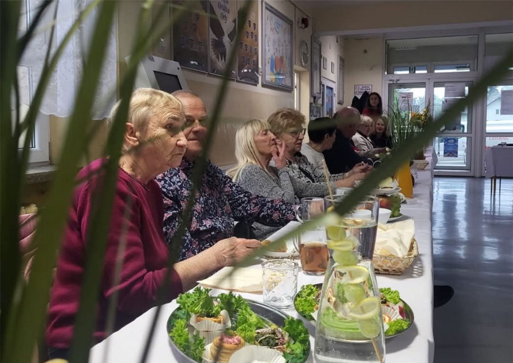 Osiem osób siedzi przy suto zastawionym stole. Na stole półmiski z wędlinami i ciastami oraz karafki z wodą i sokiem pomarańczowym.