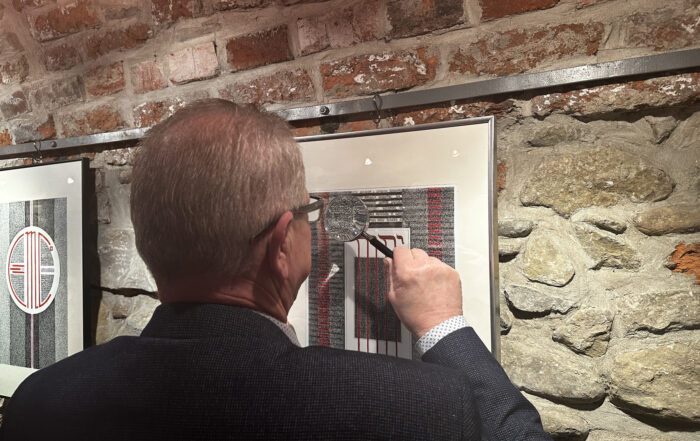 Starszy mężczyzna stoi tyłem. Trzyma w ręce lupę. Patrzy na czarno-biało-czerwoną grafikę w srebrnej ramie. Po lewej druga grafika w tej samej kolorystyce. Ramy wiszą na starej ceglanej ścianie.