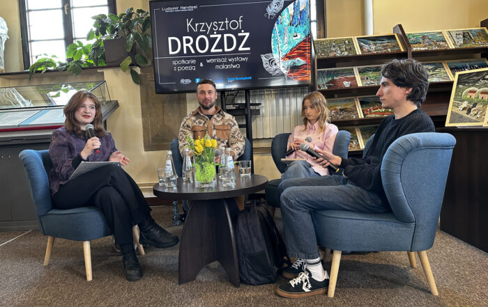 Cztery osoby siedzą na fotelach w sali przy małym okrągłym ciemnym stoliku. Troje młodych ludzi trzyma mikrofony w ręce. Czwarty mężczyzna uśmiecha. Za nimi duży ekran z grafiką z piórami. Na stole dekoracja z żółtymi tulipanami, szklane butelki i szklanki. Po prawej stronie na regałach kolorowe obrazy.