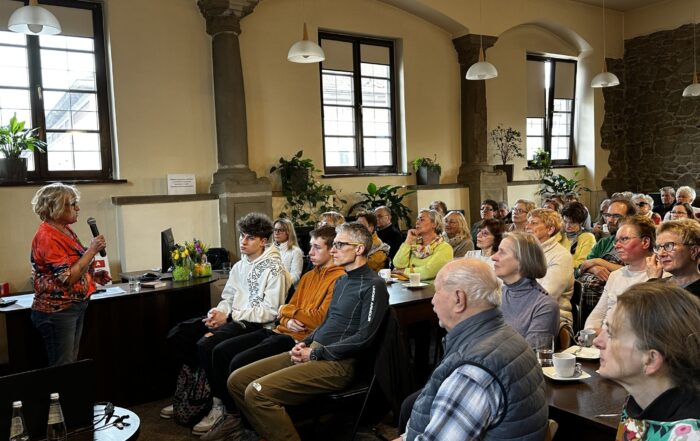 Sala z kolumnami i dużymi oknami. Po lewej stronie stoi kobieta w pomarańczowej koszulce i jeansach. Mówi do mikrofonu. Przed nią tłum ludzi. Siedzą na krzesłach i patrzą na kobietę z mikrofonem.