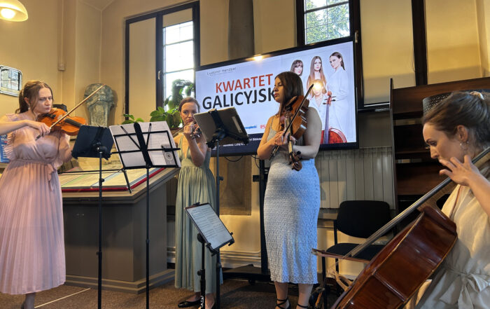 Cztery kobiety w jasnych eleganckich sukienkach. Trzy grają na skrzypcach, jedna na wiolonczeli. Za nimi w tle ekran z jasną grafiką.