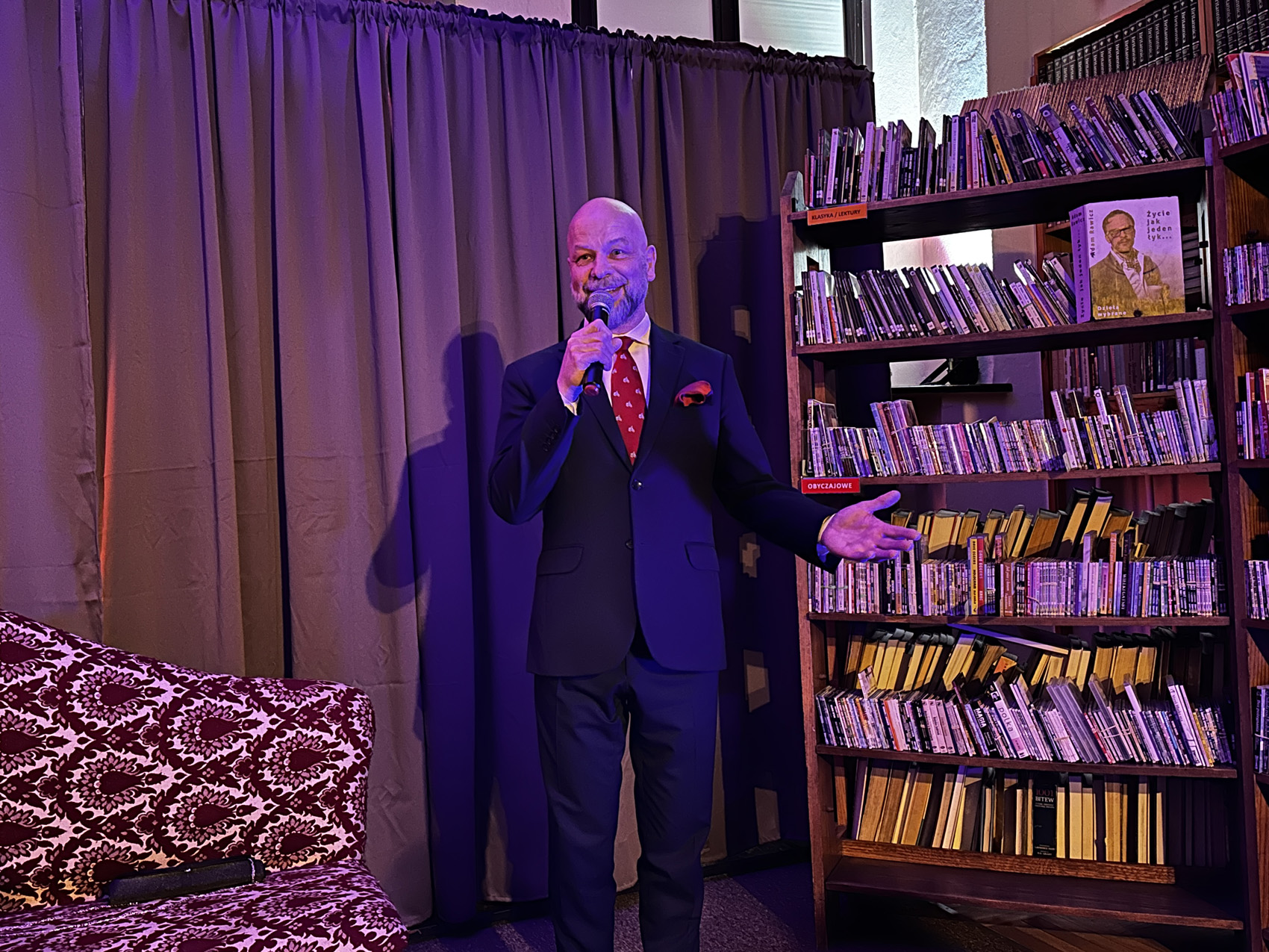 Teatr Nowy (1) Uśmiechnięty mężczyzna w czarnym garniturze i bordowym krawacie mówi do mikrofonu. W tle jasnobrązowa kotara. Po prawej stronie regały z książkami.