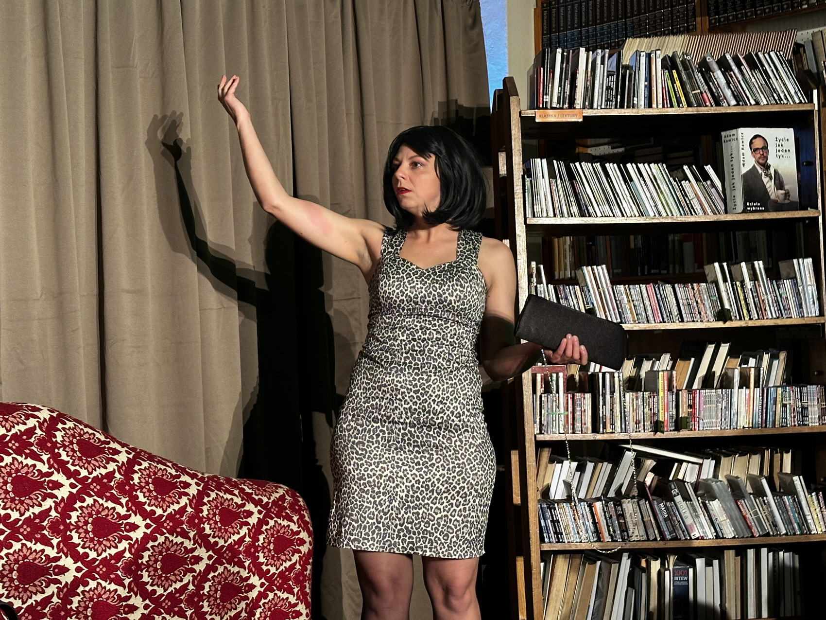 Teatr Nowy (2) Kobieta w krótkiej obcisłej sukience stoi z prawą ręką podniesioną do góry. W lewej trzyma czarną kopertową torebkę. W tle jasnobrązowa kotara. Po prawej stronie regały z książkami.