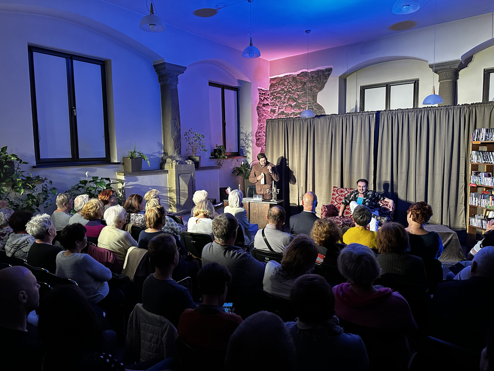 Teatr Nowy (4) Wypełniona publiką sala. Ludzie siedzą na krzesłach i patrzą scenkę przedstawianą przez dwóch mężczyzn. Jeden z nich siedzi na szezlongu w biało-czerwone wzory. Drugi stoi przy biurku i trzyma w rękach mleczną butelkę. W tle jasnobrązowa kotara. Po prawej stronie regały z książkami.