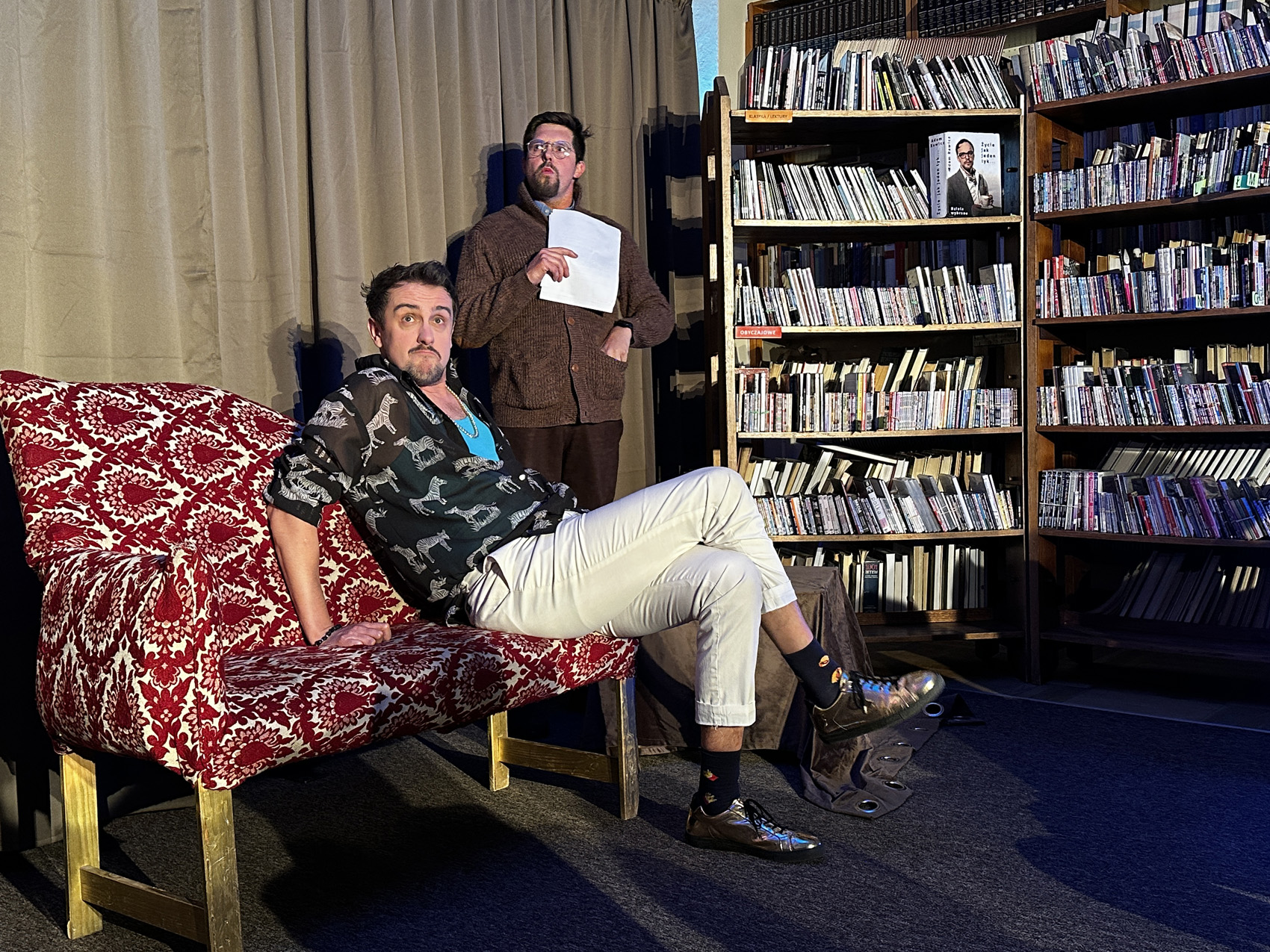 Teatr Nowy (5) W centralnym punkcie szezląg w biało-czerwone wzory. Siedzi na nim mężczyzna w jasnych spodniach i czarnej koszuli z nadrukowanymi białymi zebrami. Za nim stoi mężczyzna w brązowym swetrze i okularach, trzyma w ręku kartkę i patrzy w dal. W tle jasnobrązowa kotara. Po prawej stronie regały z książkami.