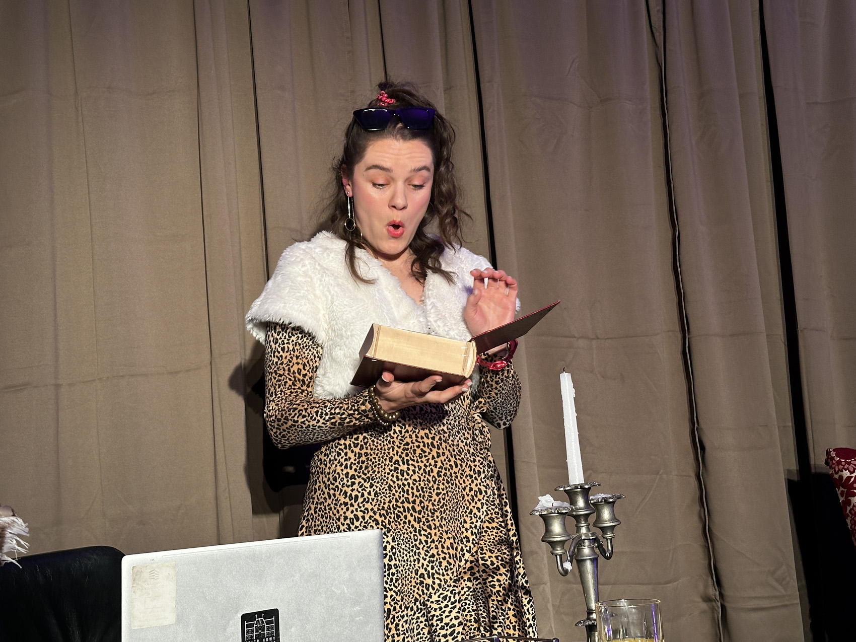 Teatr Nowy (7) Kobieta trzyma w prawej ręce otwartą książkę. Patrzy na nią zdziwiona. Ubrana jest w sukienkę w panterkowe wzory. W tle jasnobrązowa kotara.