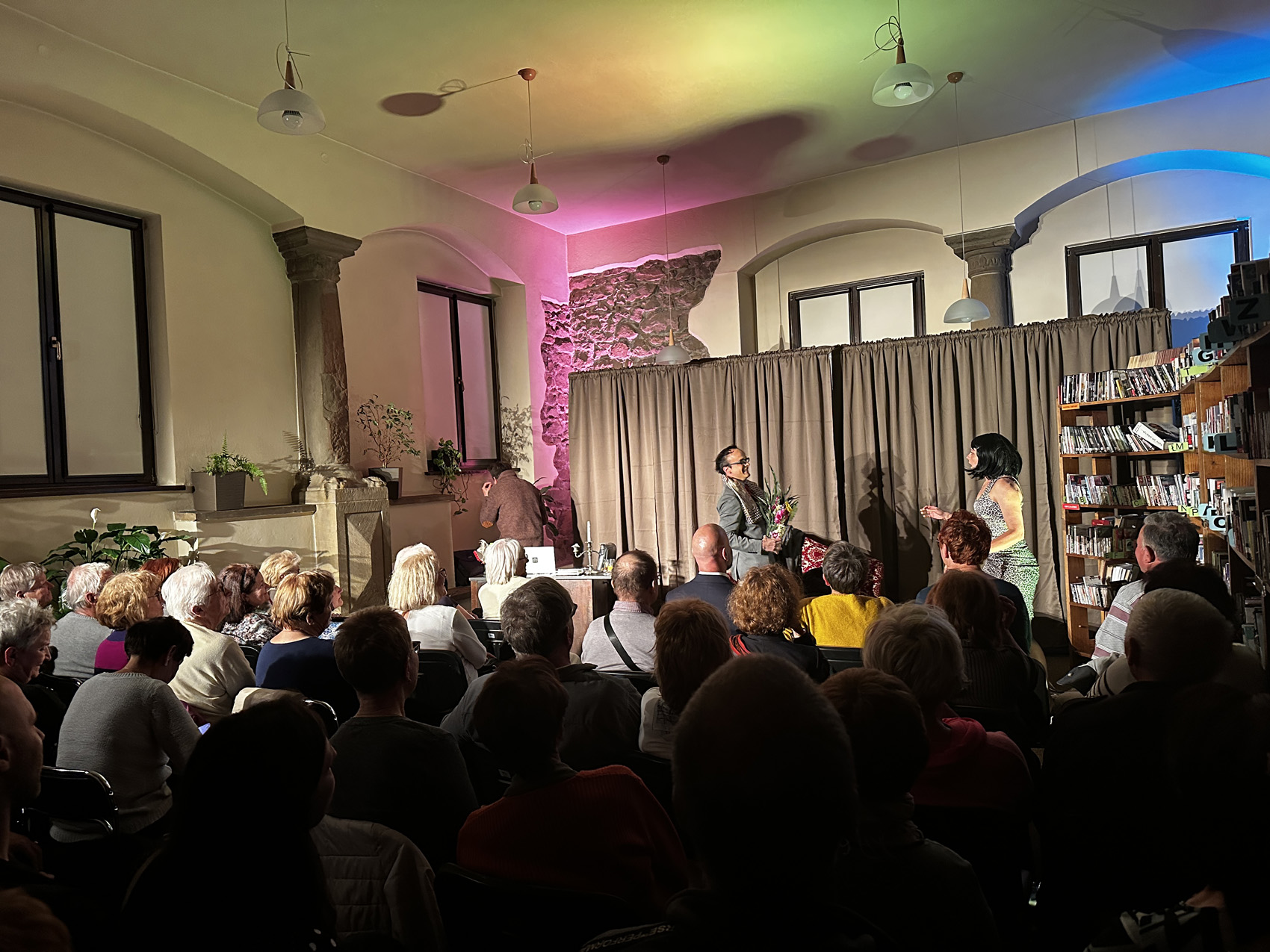Teatr Nowy (8) Wypełniona publiką sala. Ludzie siedzą na krzesłach i patrzą scenkę przedstawianą przez trzy osoby. Jeden mężczyzna stoi tyłem do publiki, drugi mężczyzna wręcza kobiecie bukiet kwiatów. W tle jasnobrązowa kotara. Po prawej stronie regały z książkami.