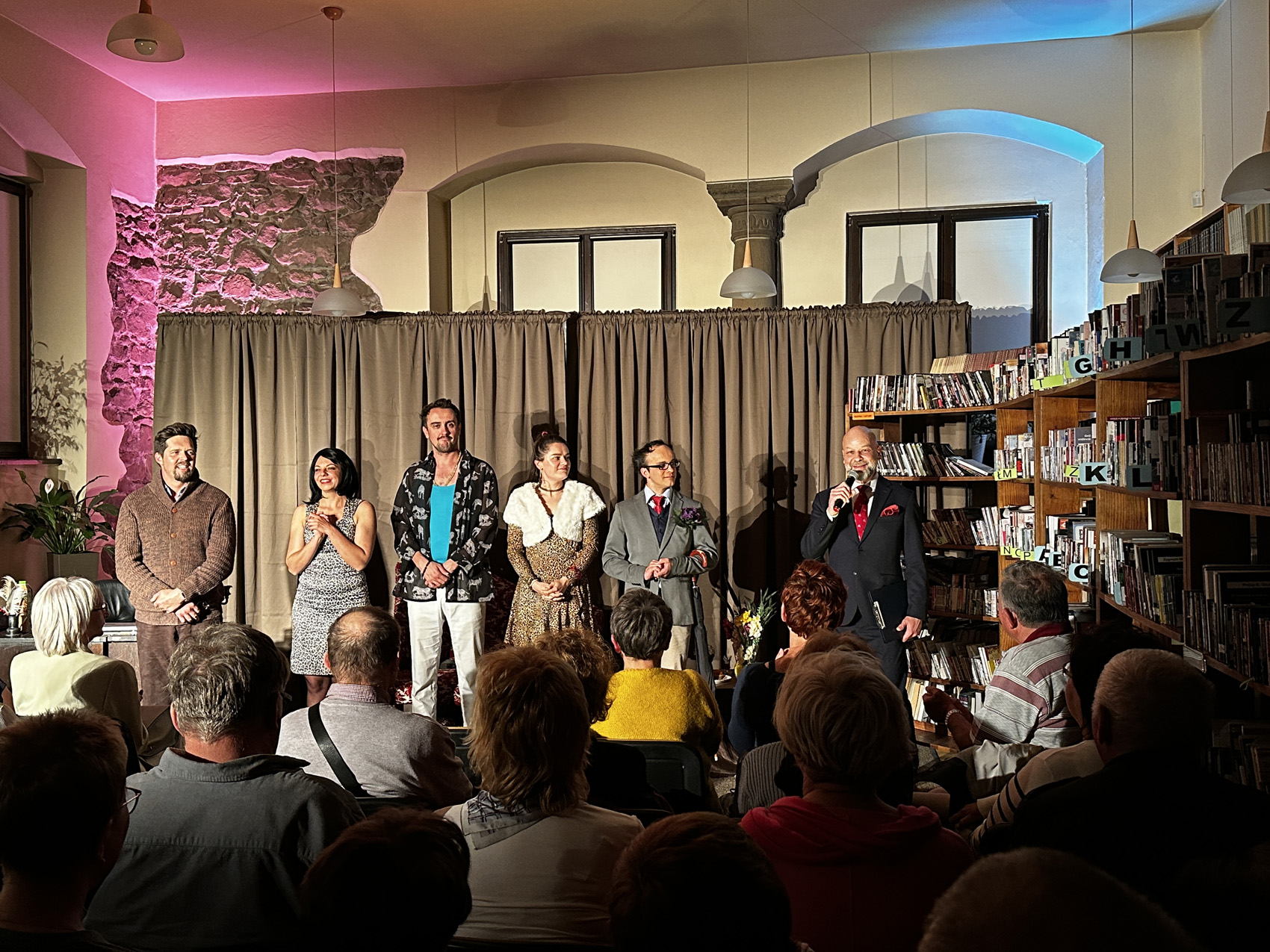 Teatr Nowy (9) Wypełniona publiką sala. Ludzie siedzą na krzesłach i patrzą na sześć osób stojących do nich frontem. Osoby uśmiechają się. Jeden mężczyzna w czarnym garniturze mówi do mikrofonu. W tle jasnobrązowa kotara. Po prawej stronie regały z książkami.