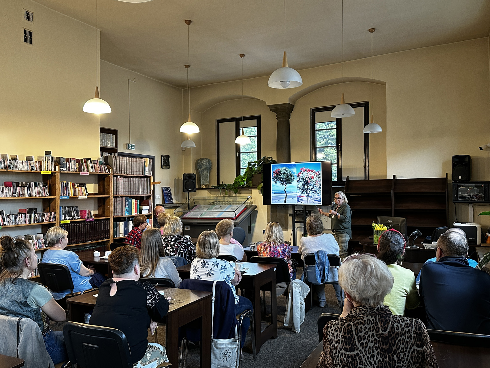 Wypełniona widownią siedzącą na krzesłach przy prostokątnych stołach sala. Na froncie ubrana na zielono kobieta ze wskaźnikiem w ręku. Kobieta pokazuje na duży ekran, na którym wyświetlone są dwa zdjęcia kolorowych drzew.