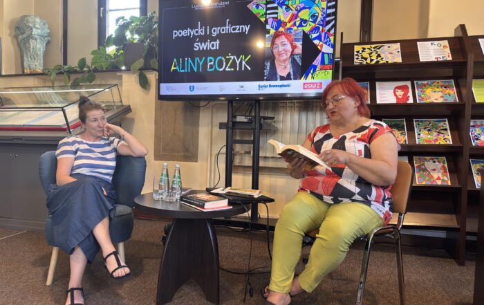 Dwie kobiety siedzą na krzesłach w sali. Kobieta po prawej ma czerwone krótkie włosy i kolorowe ubranie. Czyta książkę. Pomiędzy nimi znajduje się ciemny, okrągły, niski stolik. Na stoliku są książki, laptop i dwie butelki. Za nimi duży ekran z kolorową grafiką i połka z kolorowymi zdjęciami.