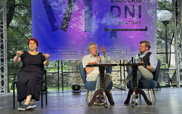 Trzy osoby siedzą na fotelach na scenie. kobieta po lewej ma czerwone włosy i czarną długą sukienkę. Gestykuluje. Obok niej siedzi dwóch mężczyzn. Trzymają mikrofony w ręce. Przed nimi są dwa małe drewniane stoliki. W tle baner podświetlony na nieskiego-fioletowo.