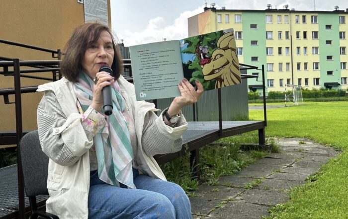 Kobieta w brązowych włosach, kolorowej apaszce i jasnej kurtce siedzi na krześle. W jednej ręce trzyma mikrofon, a w drugiej otwartą książkę z obrazkiem lwa i myszki. W tle widać metalową barierkę i żółtozielony blok.