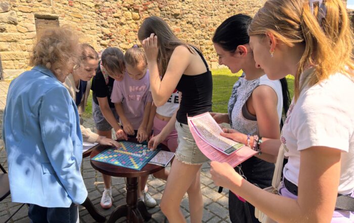 Na zdjęciu widać osiem kobiet w parku. Po prawej stronie stoi starsza kobieta w jasnych włosach i niebieskiej marynarce, która patrzy na dziewczyny przed nią, które układają litery na planszy. plansza leży na ciemnym stoliku. Po prawej stronie stoi dziewczyna w jasnych włosach, która patrzy na różową kartkę i mapę, którą trzyma w ręce. Obok niej widać dziewczynę w czarnych włosach i białej koszulce, która patrzy na planszę. W tle widać kamienne mury i zieloną trawę.