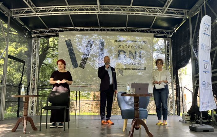 Trzy osoby na scenie w parku. Mężczyzna w ciemnym garniturze stoi w centralnej części i mówi do mikrofonu. Po bokach stoją dwie kobiety. Przed nimi znajdują się niskie stoliki i fotele. Za nimi metalowa konstrukcja sceny.