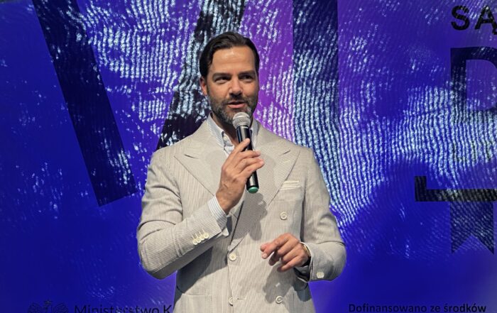 Mężczyzna o hiszpańskiej urodzie. Ubrany w jasną marynarkę. Trzyma w ręce mikrofon. Uśmiecha się. Niebiesko-fioletowe tło.