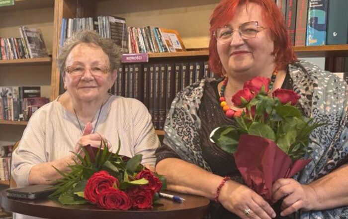 Portret dwóch kobiet. Kobiety się uśmiechają i siedzą przy okrągłym brązowym stoliku. Przed jedną z kobiet leży bukiet bordowych róż, druga kobieta trzyma bukiet w rękach.