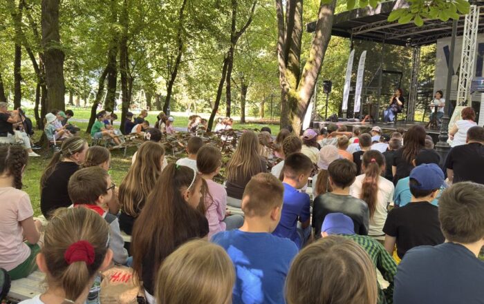 Tłum ludzi siedzi na ławkach w parku. Przed nimi scena. Na scenie w fotelach siedzą dwie kobiety. W tle park i dużo drzew.