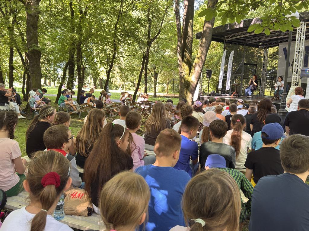 Tłum ludzi siedzi na ławkach w parku. Przed nimi scena. Na scenie w fotelach siedzą dwie kobiety. W tle park i dużo drzew.